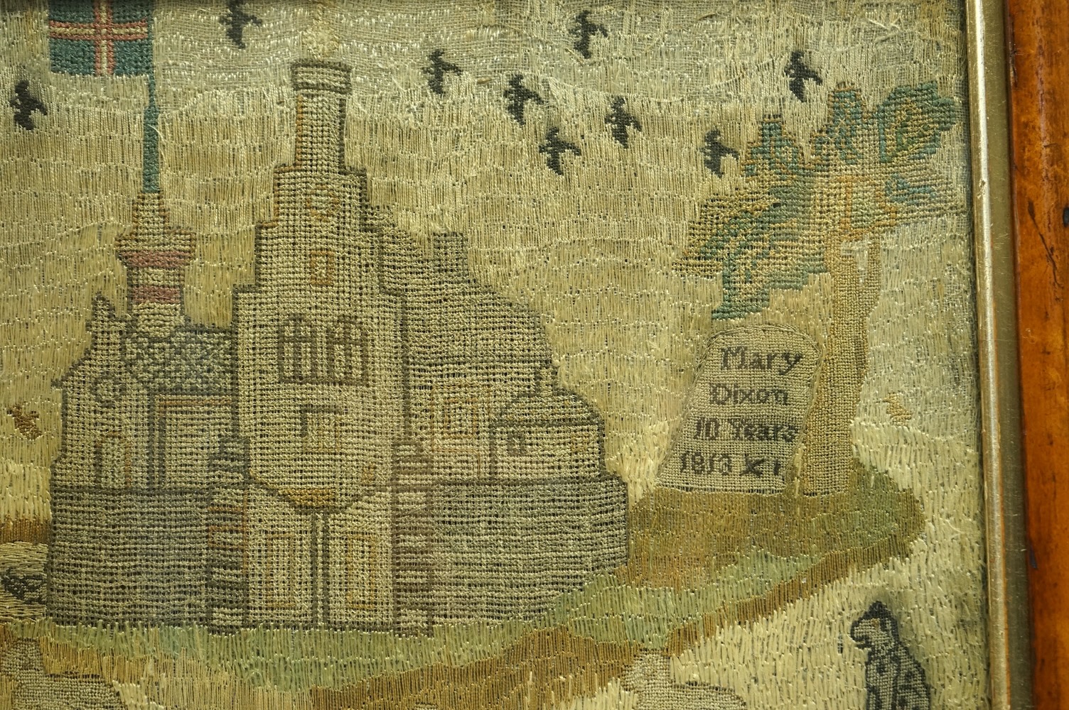 An unusual 19th century maple framed embroidery, in the form of a country scene embroidered with sheep, birds, ducks and a dog amongst houses and a church. The single grave stone signed Mary Dixon, 10 years, 1813. 30.5cm
